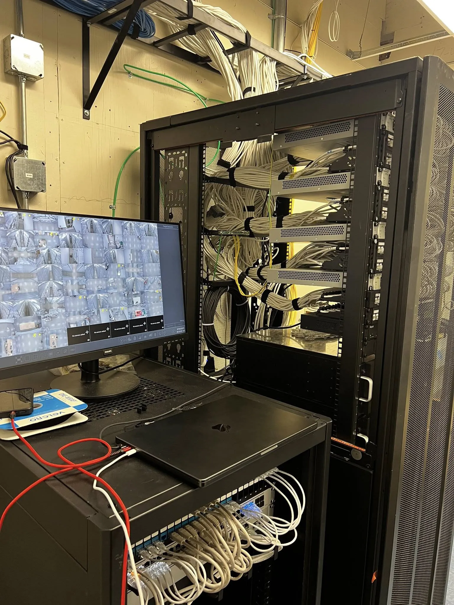 Open server rack in a data closet with neatly bundled network cables and a monitor displaying a security camera grid