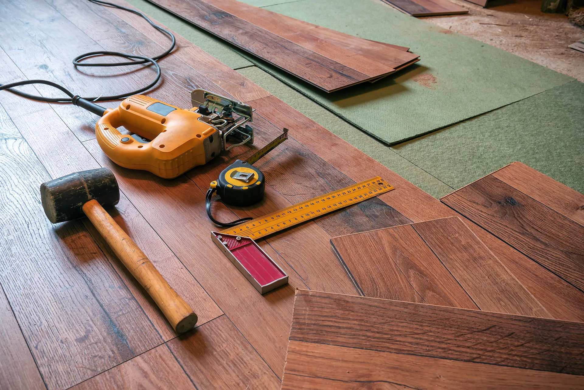 Laminate flooring installation tools, including a jigsaw, tape measure, square, and rubber mallet, resting on newly laid planks and green underlayment.