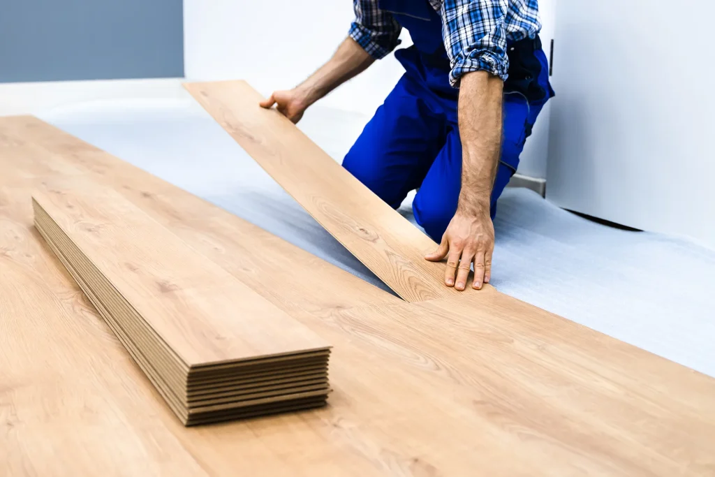 A worker in blue overalls installing light oak laminate wood flooring planks in a room renovation project.